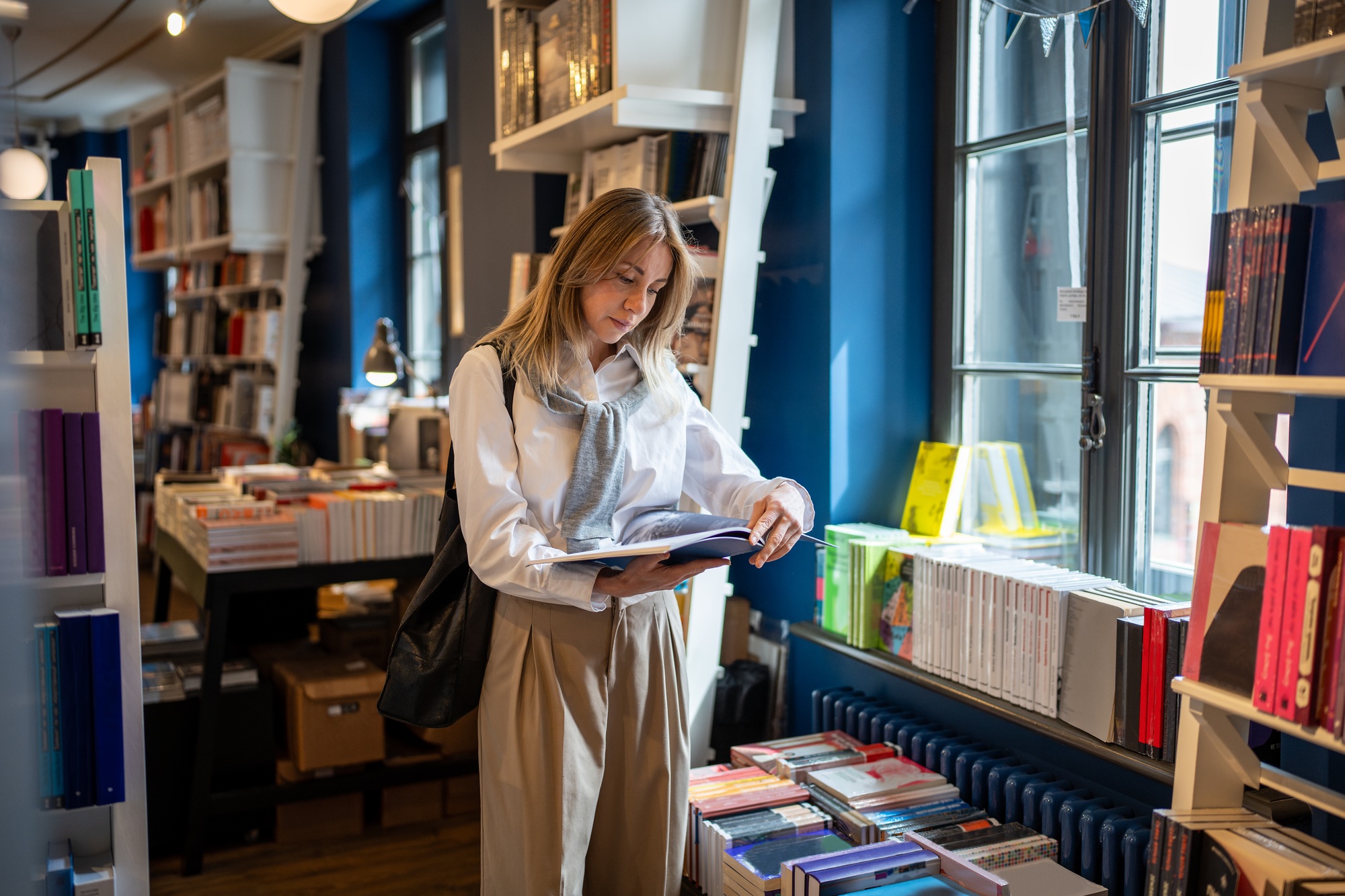 Focused thoughtful middle aged woman reading book standing shopping in modern bookstore near window